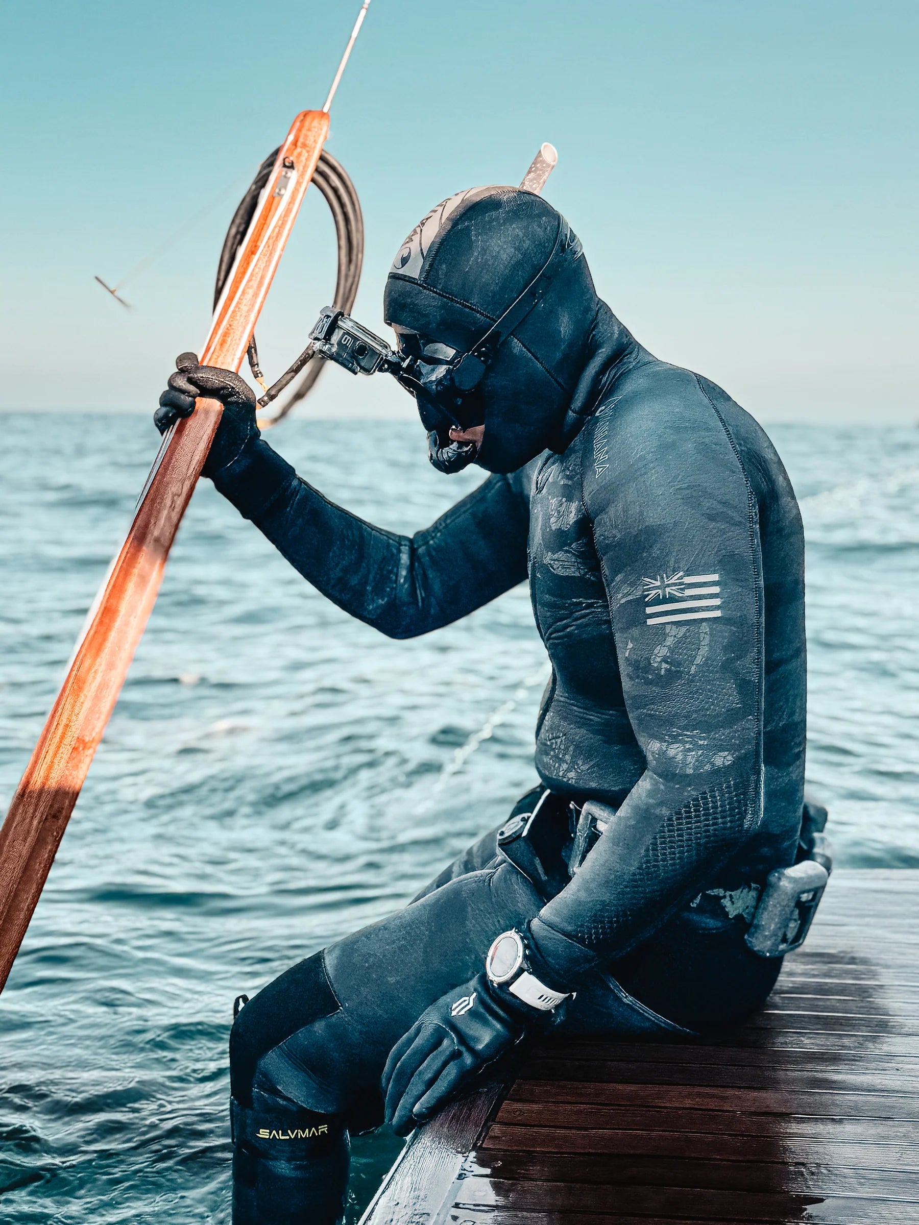 Person in a wetsuit sitting on a dock with fishing equipment, water in the background